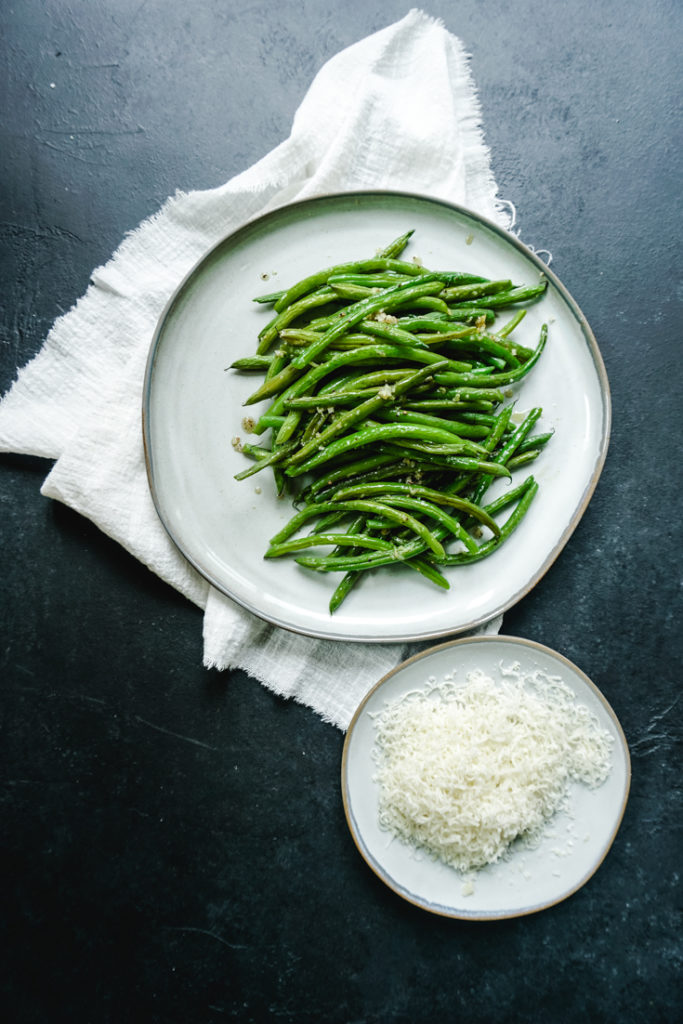 Baked green beans piled up on a plate with a white napkin under it and a smaller plate of grated parmesan next to it