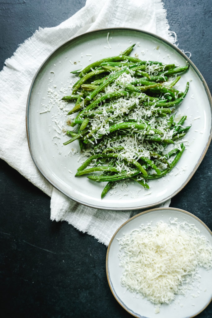 Green beans piled on a plate with grated parmesan on top and a plate of grated parmesan on the side