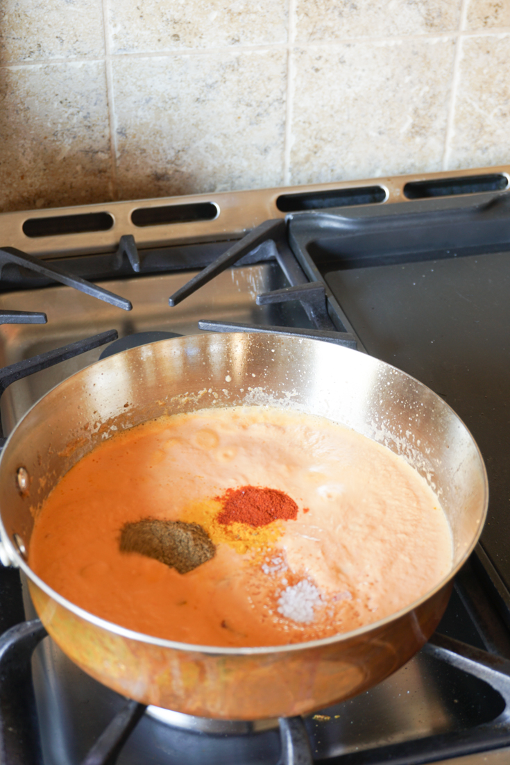 Stainless steel sauce pan on stove with sauce in it and spices on top of it