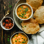 Top wide shot of a platter with a bowl of potato curry and three pooris and another bowl of potato curry and a bowl of pickle to the side of the platter