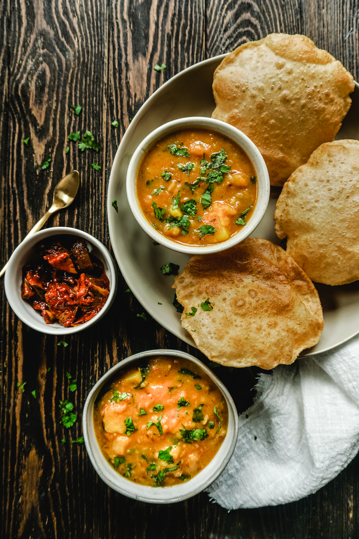 Top wide shot of a platter with a bowl of potato curry and three pooris and another bowl of potato curry and a bowl of pickle to the side of the platter