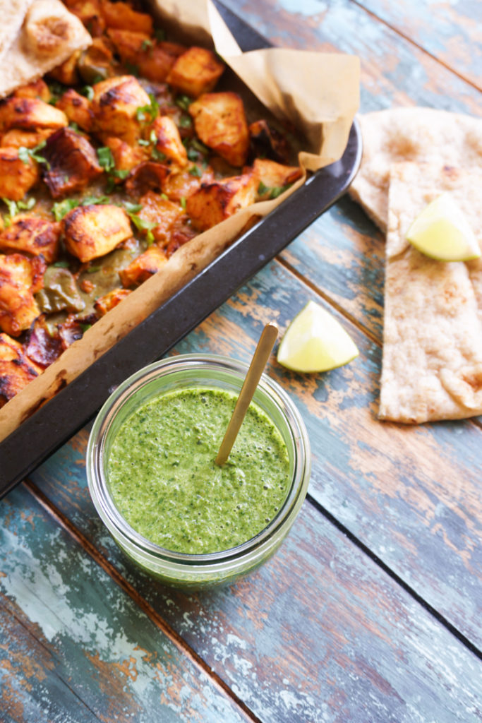 Jar of cilantro mint chutney next to a tray of delicious food and some flatbread next to it