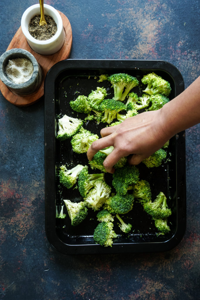 A hand tossing broccoli in a black sheet pan