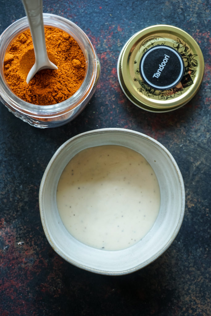 A small bowl of ranch and a glass storage bottle with tandoori spice in it with a white spoon sticking out of it