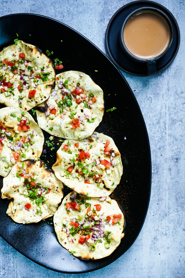 Masala papads placed in a large black platter with a black up of chai next to it