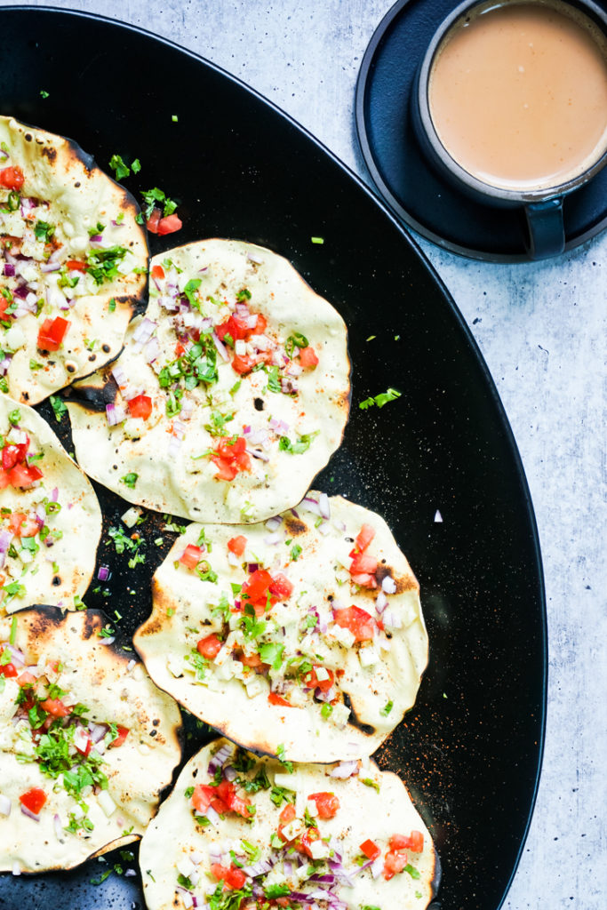 Masala papads placed in a large black platter with a black up of chai next to it