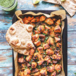 Sheet pan with chicken tikka and vegetables with two naans on top of it and a naan on the side outside