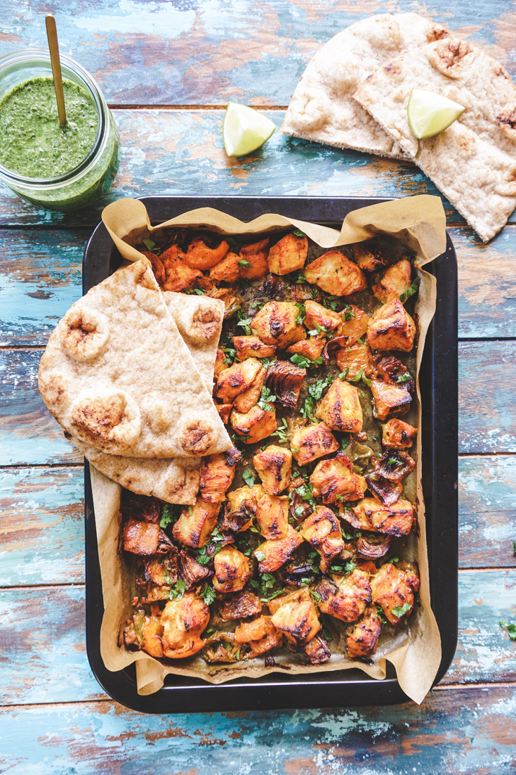 Sheet pan with chicken tikka and vegetables with two naans on top of it and a naan on the side outside