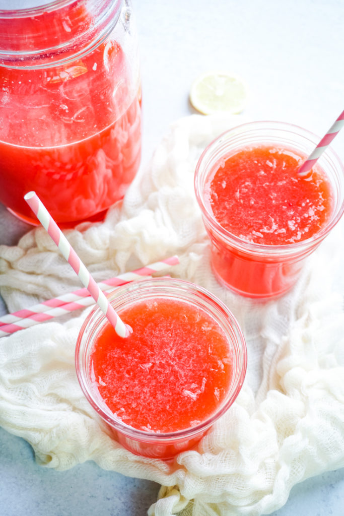 Two glasses of strawberry lemonade with pink straws in them next to a pitcher with lemonade