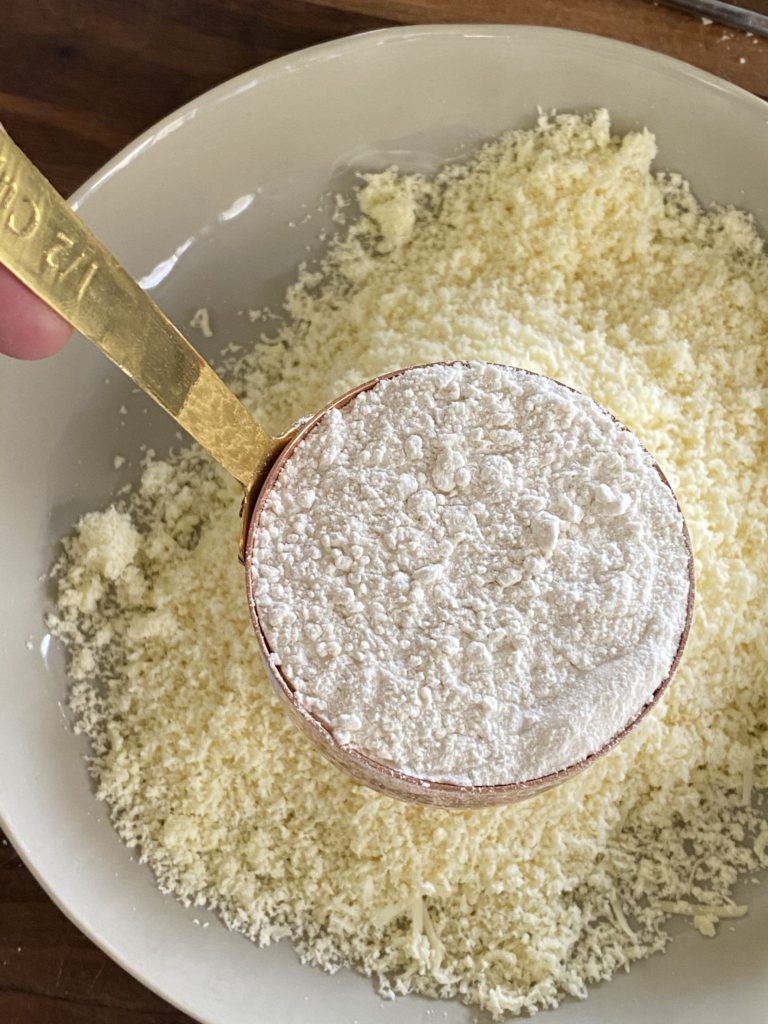 measuring cup of all purpose flour held on top of a bowl of grated khoya