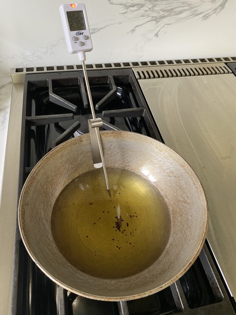 a kadhai on stove with oil in it and a candy thermometer plugged in