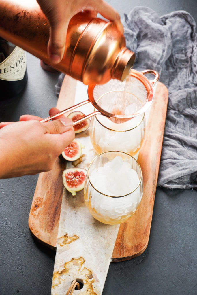 Two glasses on a paddle board and cocktail being poured into one through a tea strainer