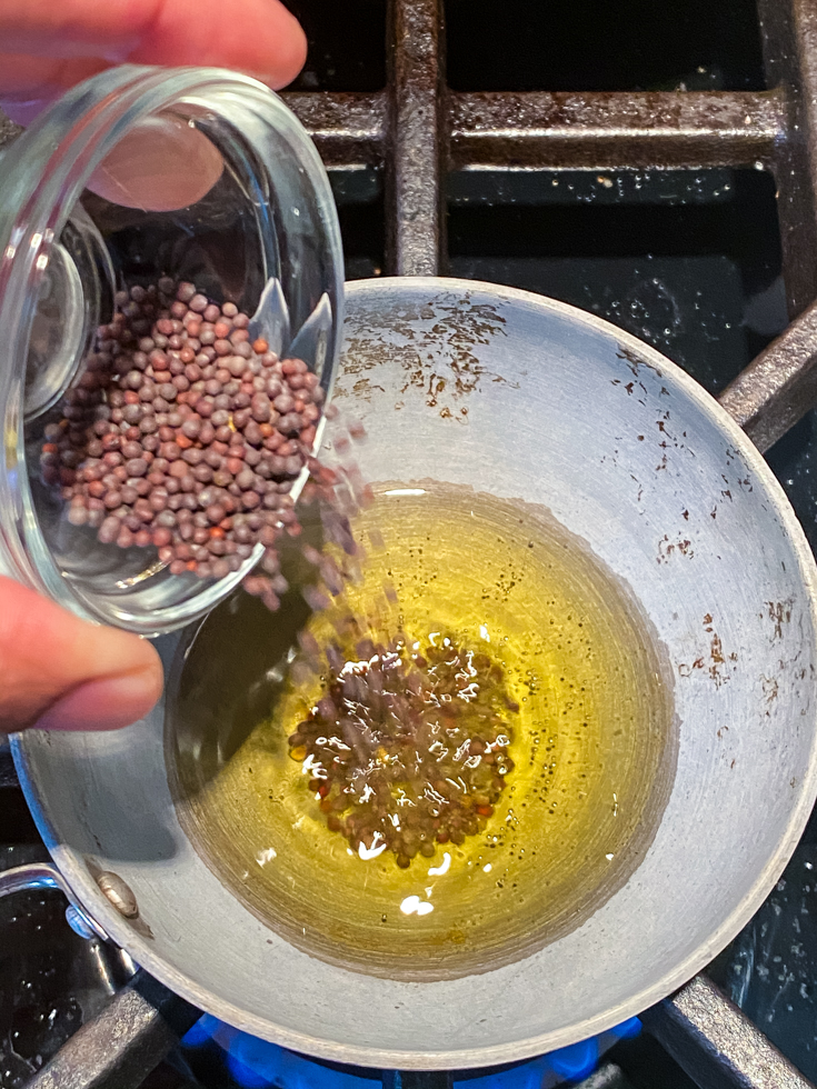 small pan with oil in it and a hand adding mustard seeds to it