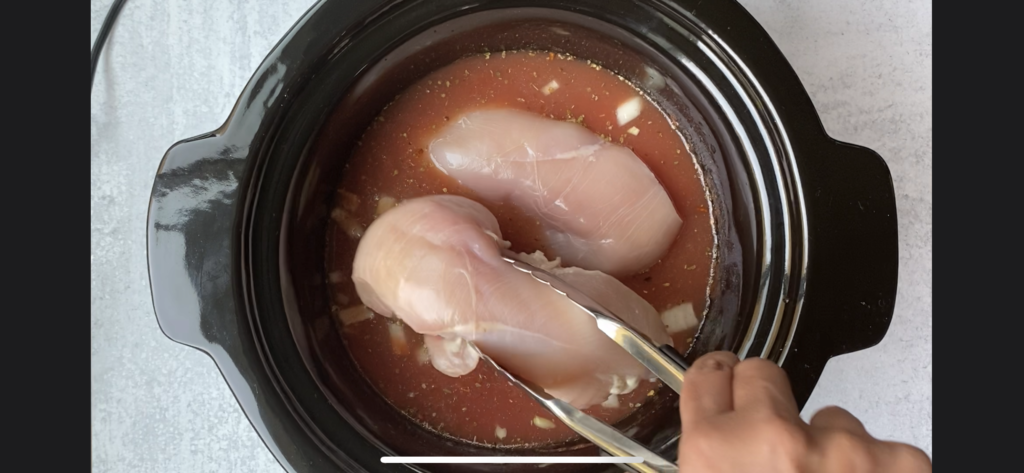 hand placing chicken breast inside crockpot with a pair of tongs