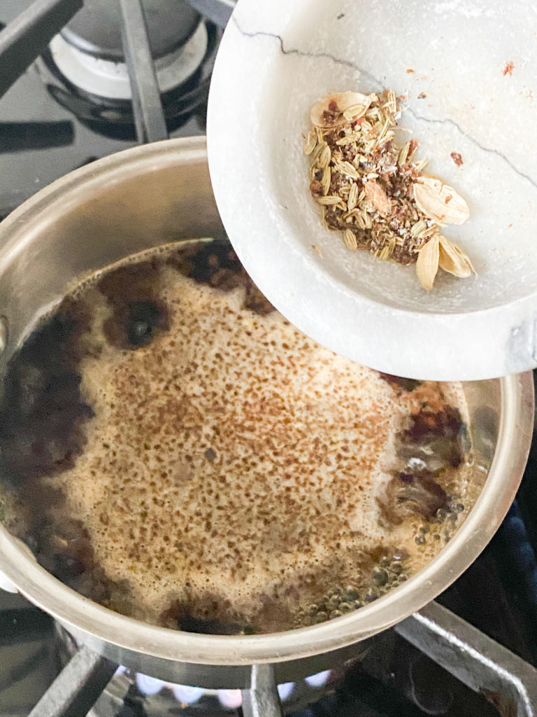 spices being added to a saucepan with boiling water and tea leaves in it