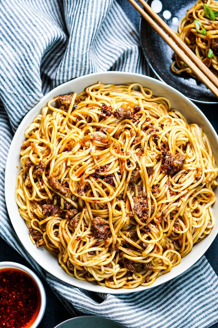 spicy pork noodles in a white bowl with two small bowl of chili oil and scallions next to it