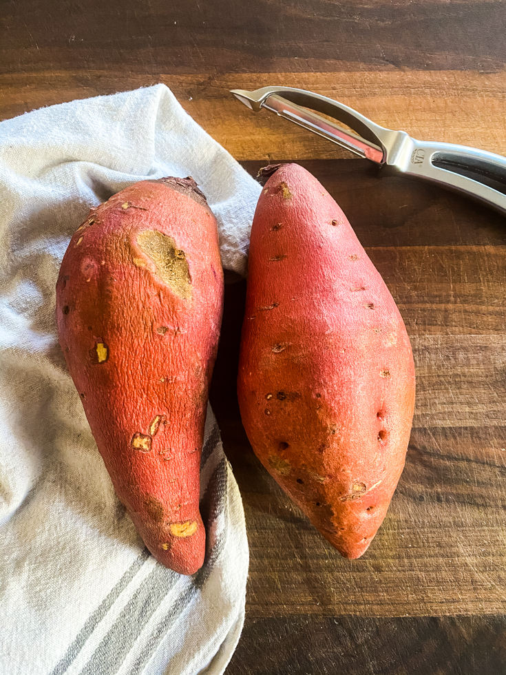 two sweet potatoes and a peeler on a chopping board