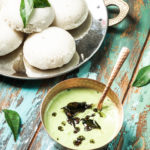 a copper bowl of cilantro coconut chutney with a plate of idlis on the side