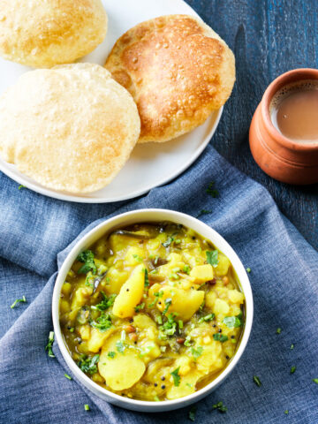 potato curry in a bowl and three pooris in a plate with a cup of chai next to it