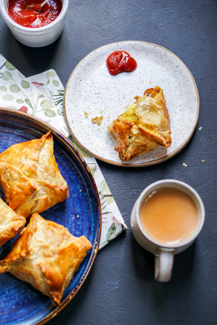 A cup of chai with an egg puff and ketchup on a plate next to it with a blue plate of egg puffs in it as well