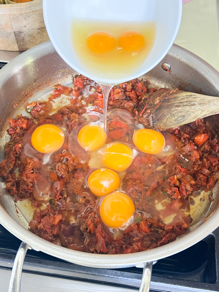 eggs being added from a white bowl to the onion tomato mixture in a pan on the stove