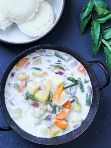 Stew with potato, carrot and green beans in coconut milk in a black kadai bowl and a bowl of idlis