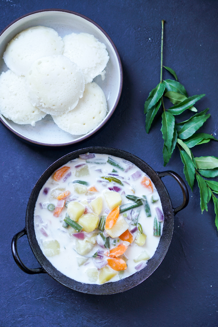 Stew with potato, carrot and green beans in coconut milk in a black kadai bowl and a bowl of idlis