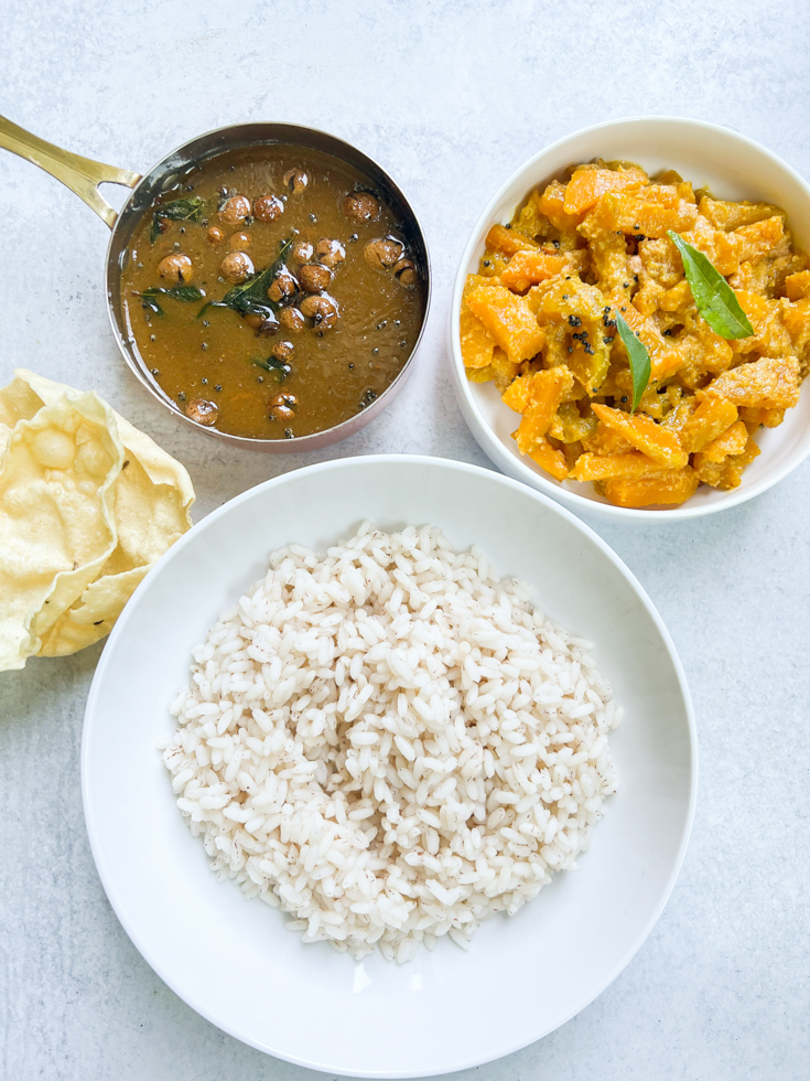 a white bowl of rice, a smaller white bowl of pumpkin curry and a pan of vathal kuzhambu next to it