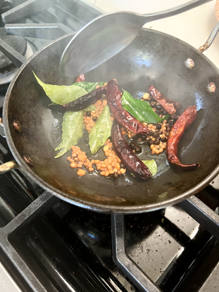 curry leaves, chillies and lentils in a black pan being stirred by a spoon