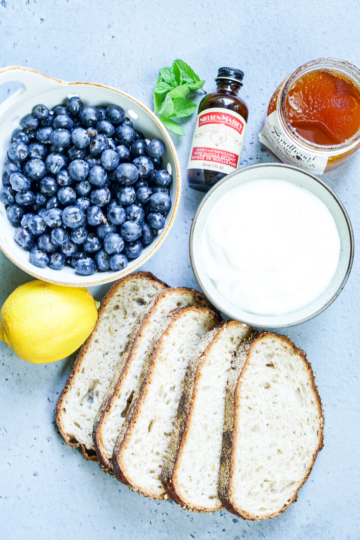 all ingredients laid out - a bowl of blueberries, fresh min, bottle of vanilla, bottle of honey, bowl of yogurt, bread slices and a lemon
