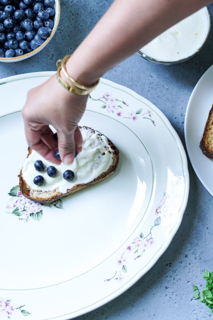 blueberries being placed on Greek Yogurt and Blueberry topped toast on a white platter
