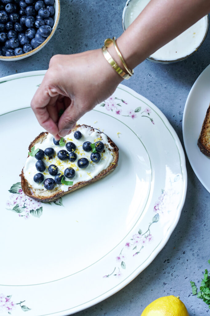 hand placing mint leaves on Greek Yogurt and Blueberry topped toast on a white platter