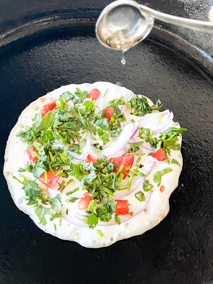oil being drizzled by a spoon on to the uthappam