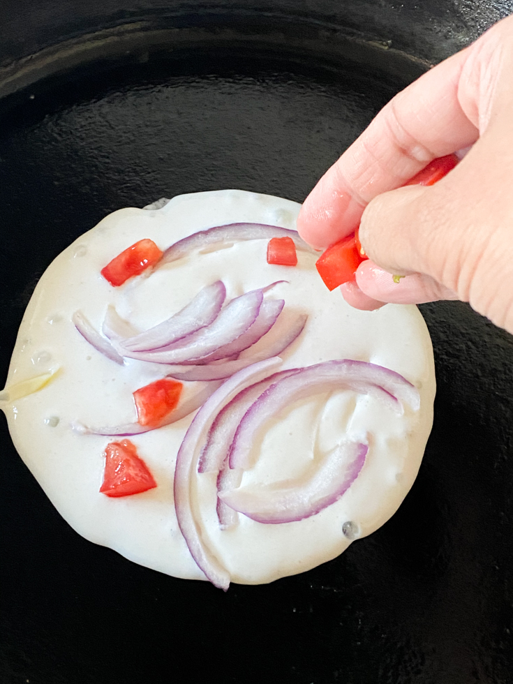 hand adding chopped tomatoes to uthappam batter in a pan